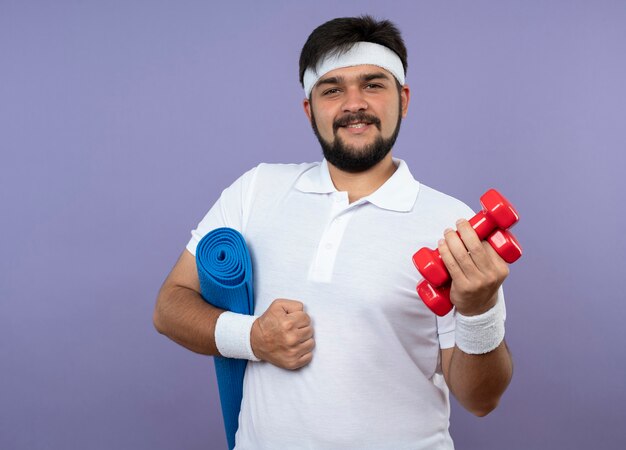 Pleased young sporty man wearing headband and wristband holding dumbbells with yoga mat