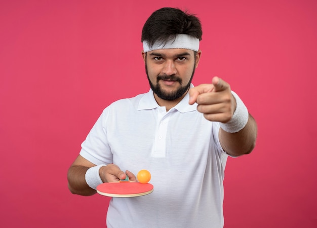 Pleased young sporty man wearing headband and wristband holding ping pong racket with ball showing you gesture