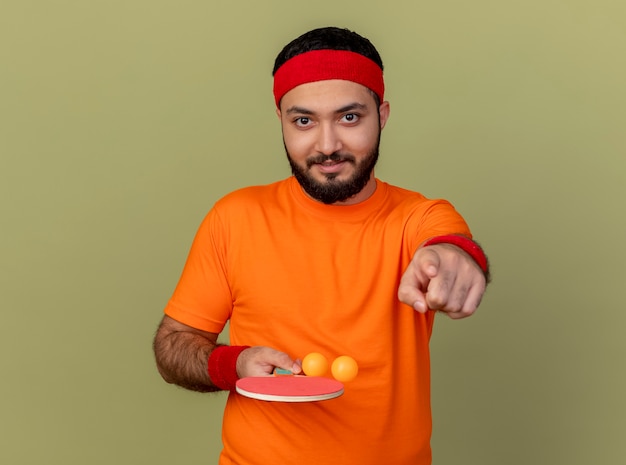 Pleased young sporty man wearing headband and wristband holding ping pong racket with balls and showing you gesture isolated on olive green background