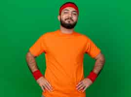 Free photo pleased young sporty man wearing headband and wristband putting hands on hip isolated on green background