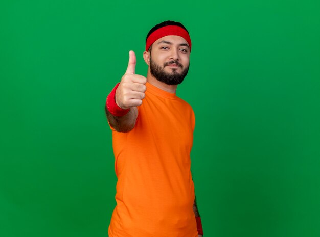 Pleased young sporty man wearing headband and wristband showing thumb up isolated on green