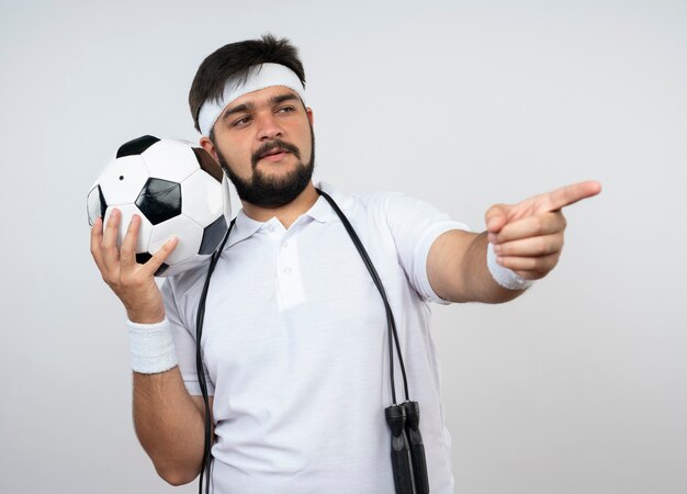 Pleased young sporty man wearing headband and wristband with jump rope on shoulder holding ball points at side isolated on white wall