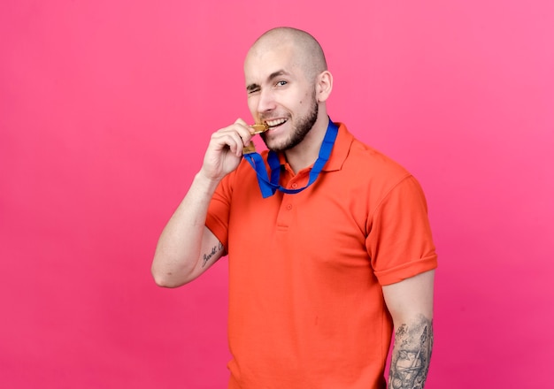 Pleased young sporty man wearing and holding medal in mouth isolated on pink wall