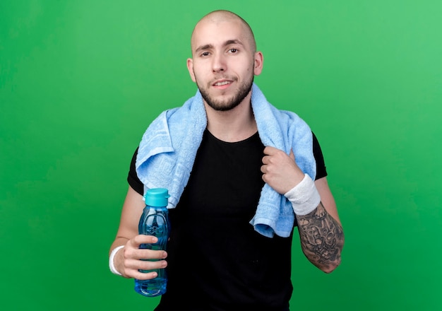 Pleased young sporty man wearing wristband holding water bottle with towel on shoulder isolated on green wall