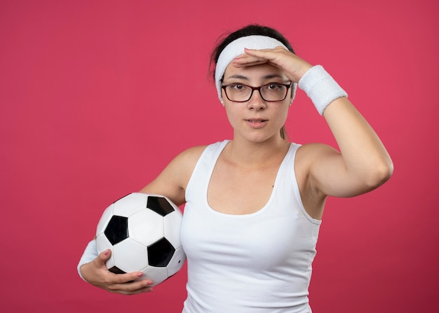 Pleased young sporty woman in optical glasses wearing headband and wristbands keeps palm at forehead and holds ball isolated on pink wall
