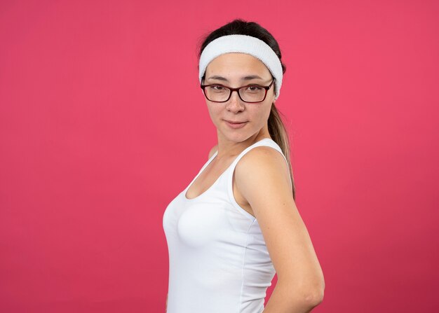 Довольная молодая спортивная женщина в оптических очках с повязкой на голову и браслетами стоит боком, изолированным на розовой стене