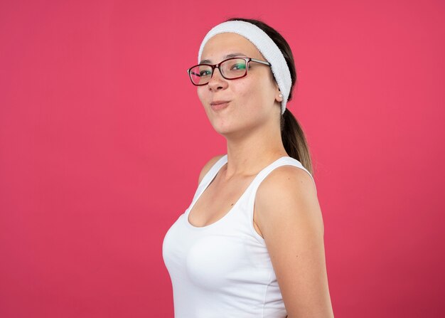 Pleased young sporty woman in optical glasses wearing headband and wristbands stands sideways and looks at front isolated on pink wall