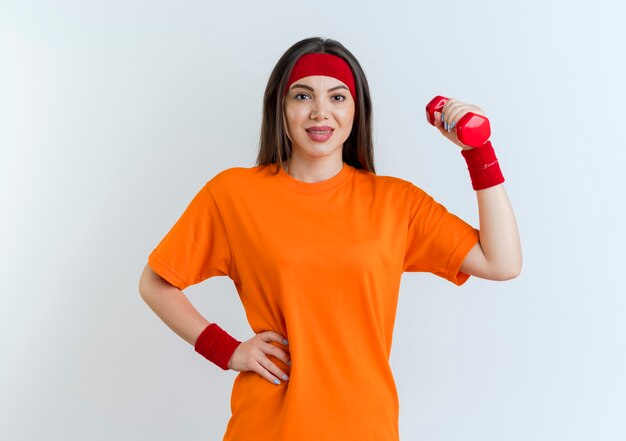 Pleased young sporty woman wearing headband and wristbands holding dumbbell  keeping hand on waist isolated on white wall with copy space