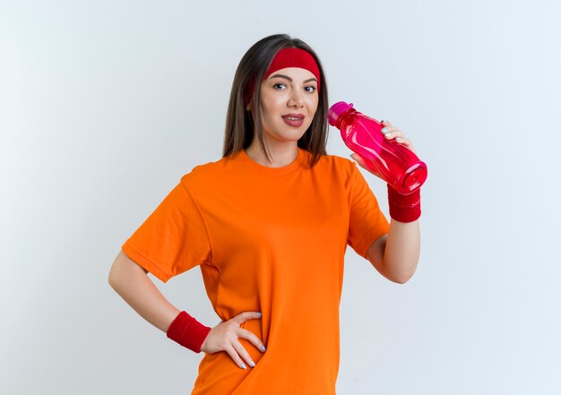 Pleased young sporty woman wearing headband and wristbands holding water bottle keeping hand on waist  isolated on white wall with copy space