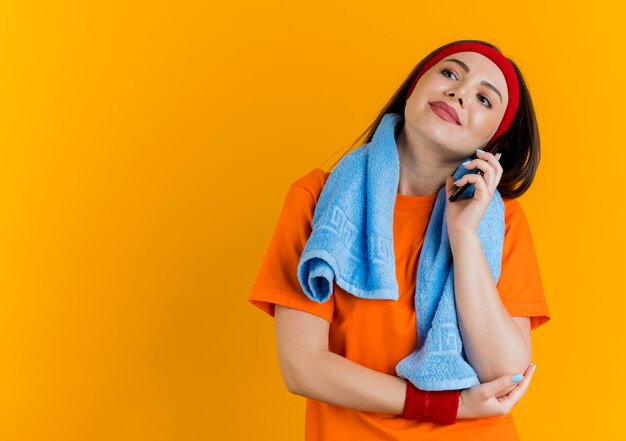 Pleased young sporty woman wearing headband and wristbands with towel around neck looking at side putting hand on elbow talking on phone 