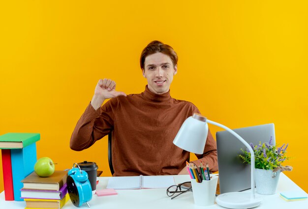 Pleased young student boy sitting at desk with school tools his thumb down on yellow