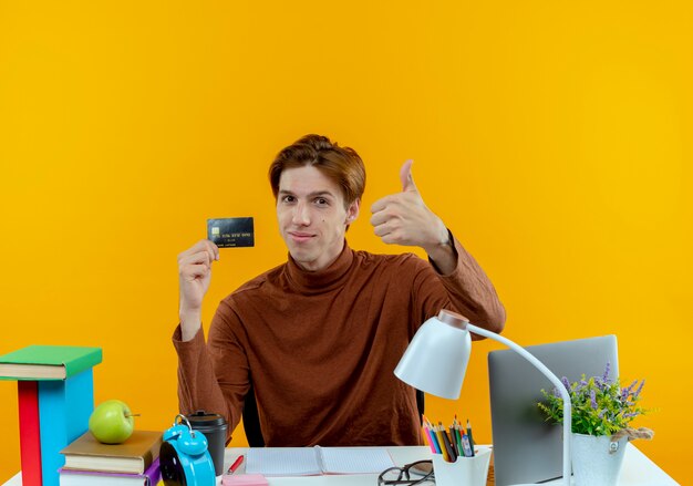 pleased young student boy sitting at desk with school tools holding credit card his thumb up