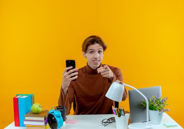 Pleased young student boy sitting at desk with school tools holding phone and showing you gesture isolated on yellow wall