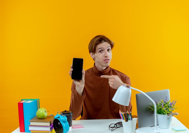Довольный молодой студент мальчик сидит за столом со школьными инструментами и указывает на телефон, изолированный на желтой стене