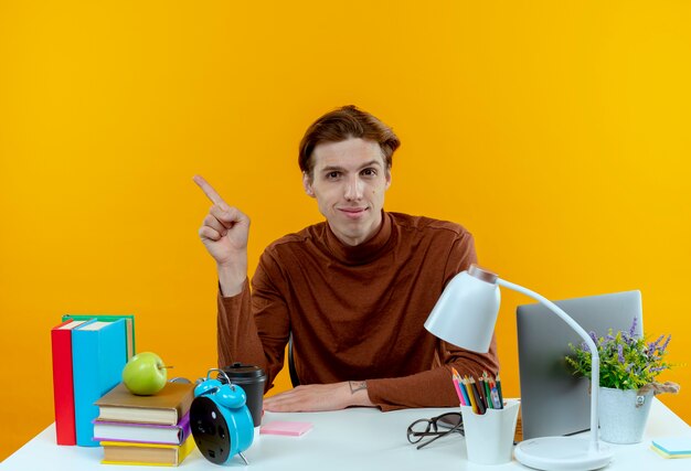 Pleased young student boy sitting at desk with school tools points at side on yellow