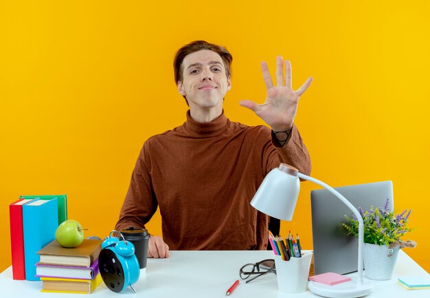 Pleased young student boy sitting at desk with school tools showing five isolated on yellow wall