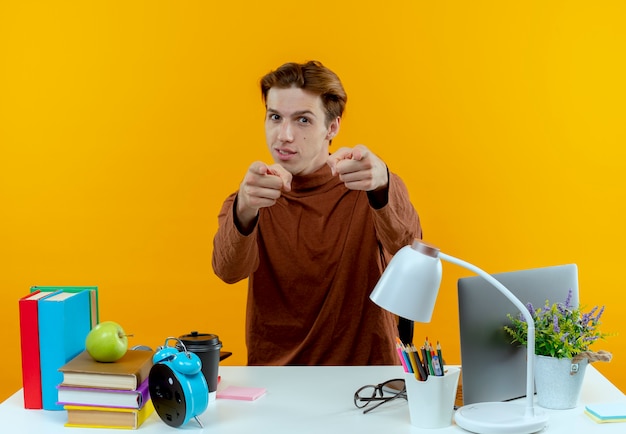 Pleased young student boy sitting at desk with school tools showing you gesture isolated on yellow wall