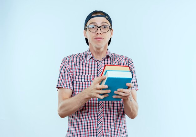 Pleased young student boy wearing back bag and glasses and cap holding books on white