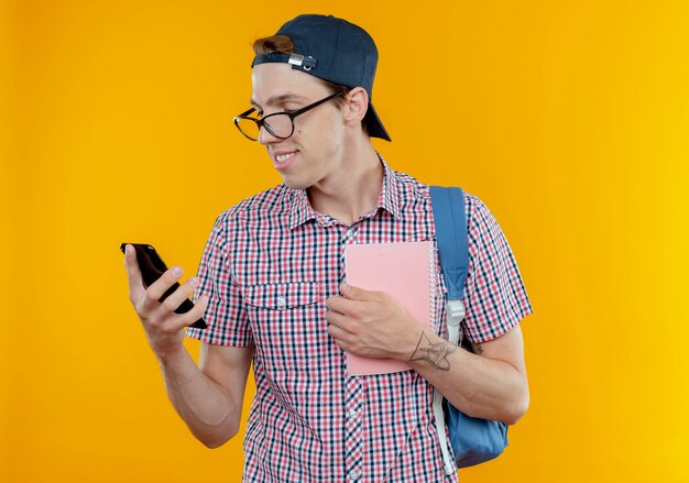 バックバッグとメガネとノートを保持し、白で隔離の彼の手で携帯電話を見てキャップを身に着けている若い学生の少年を喜ばせる
