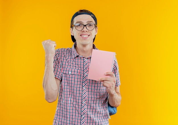Pleased young student boy wearing back bag and glasses and cap holding notebook and showing yes gesture
