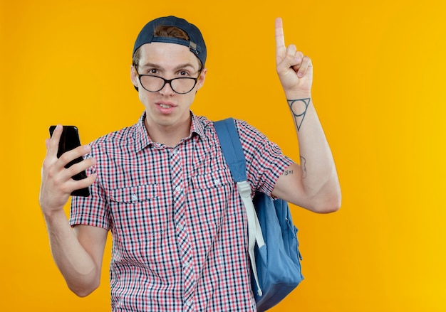 pleased young student boy wearing back bag and glasses and cap holding phone and points at up