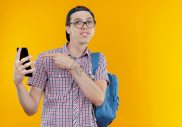 Pleased young student boy wearing back bag and glasses and cap holding and points at phone on white