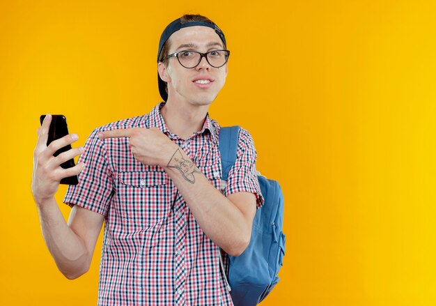 Довольный молодой студент мальчик в задней сумке и очках и кепке держит и указывает на телефон на белом
