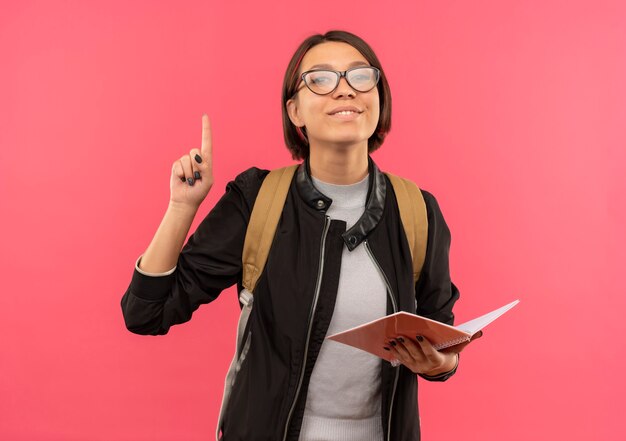 Pleased young student girl wearing glasses and back bag holding note pad raising finger isolated on pink wall