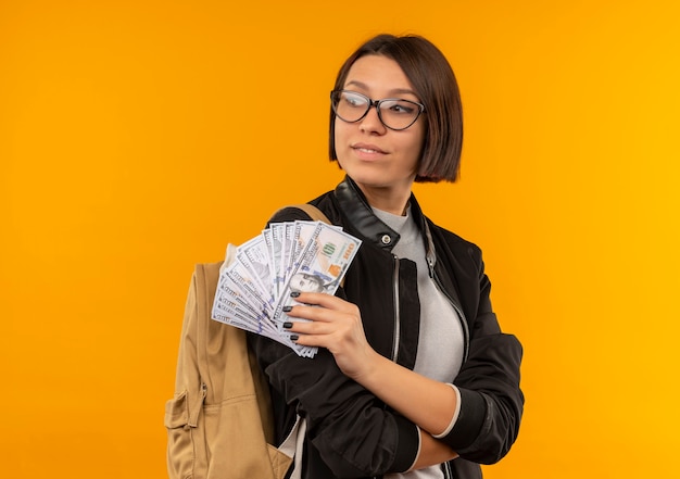 Pleased young student girl wearing glasses and back bag standing with closed posture holding money looking at side isolated on orange wall