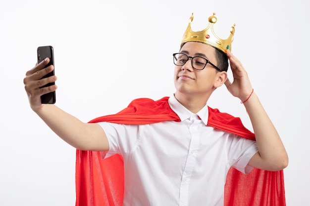 Free photo pleased young superhero boy in red cape wearing glasses and crown touching crown taking selfie isolated on white background
