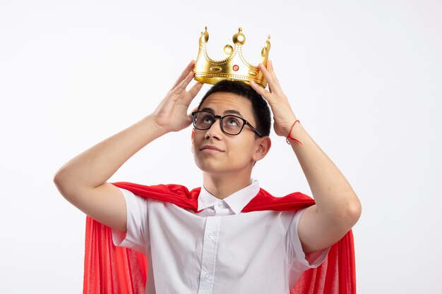 Pleased young superhero boy in red cape wearing glasses holding crown above head looking up isolated on white background