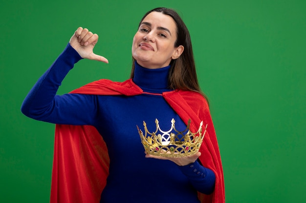 Pleased young superhero girl holding crown showing thumb down isolated on green