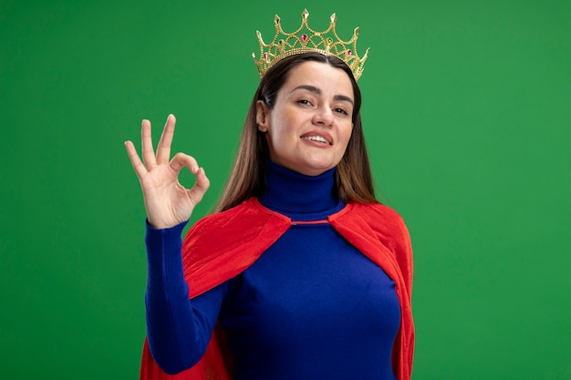 Довольная молодая девушка-супергерой в короне показывает нормальный жест, изолированный на зеленом