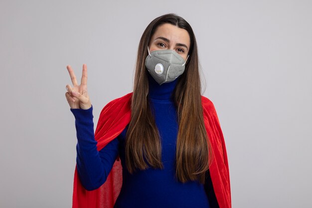Lieta giovane ragazza del supereroe che indossa la mascherina medica che mostra il gesto di pace isolato su bianco