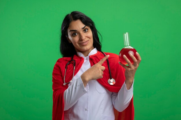 Pleased young superhero girl wearing medical robe with stethoscope holding and points at chemistry glass bottle filled with red liquid isolated on green