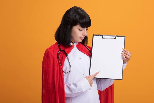 Pleased young superhero girl wearing stethoscope with medical robe and cloak holding and points at clipboard isolated on orange wall