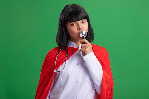 Pleased young superhero girl wearing stethoscope with medical robe and cloak kissing stethoscope isolated on green wall