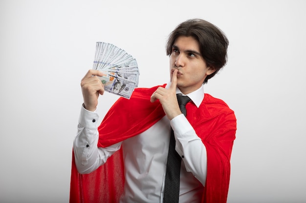 Pleased young superhero guy looking at side wearing tie holding cash and showing silence gesture