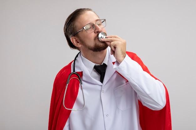 Pleased young superhero guy wearing medical robe with stethoscope and glasses kissing stethoscope isolated on white background