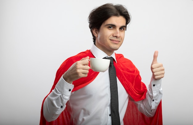 Pleased young superhero guy wearing tie holding cup of tea and showing thumb up isolated on white