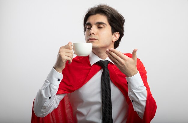 Довольный молодой супергерой с закрытыми глазами в галстуке, нюхающий кофе, изолированный на белом