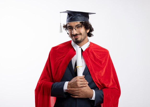 Pleased young superhero man in optical glasses wearing suit with red cloak and graduation cap holds diploma and looks at front isolated on white wall