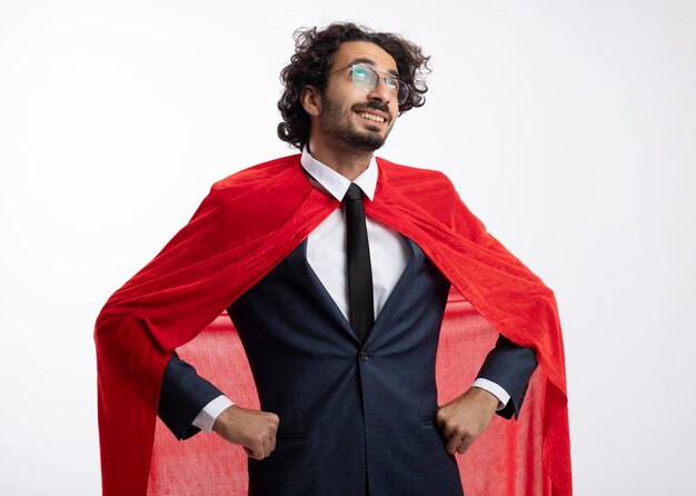 Pleased young superhero man in optical glasses wearing suit with red cloak puts hands on waist and looks up isolated on white wall