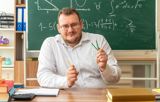 pleased young teacher wearing glasses sitting at desk with school supplies in classroom holding counting sticks looking at front