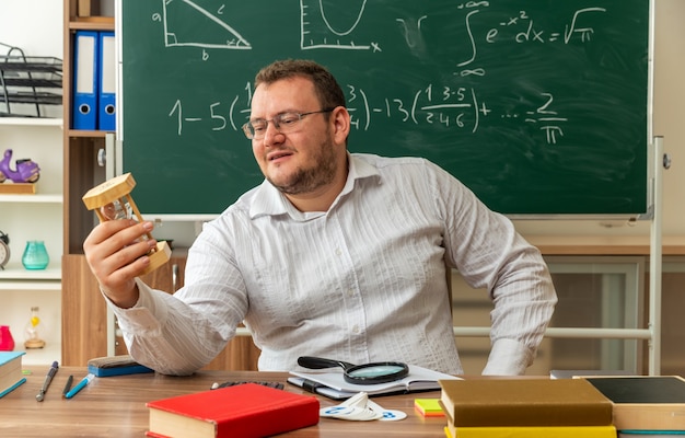 Foto gratuita compiaciuto giovane insegnante con gli occhiali seduto alla scrivania con materiale scolastico in classe tenendo la mano sulla vita tenendo e guardando la clessidra