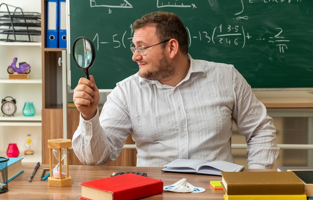 довольный молодой учитель в очках сидит за столом со школьными принадлежностями в классе, глядя в сторону через увеличительное стекло