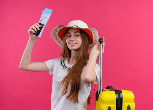 Довольная молодая путешественница в шляпе держит кредитную карту, билеты на самолет и чемодан на изолированном розовом пространстве