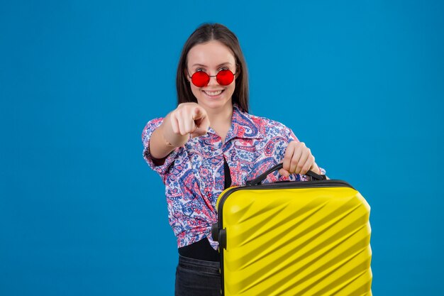 Pleased young traveler woman wearing red sunglasses holding yellow suitcase pointing with finger to camera smiling cheerfully standing over blue background