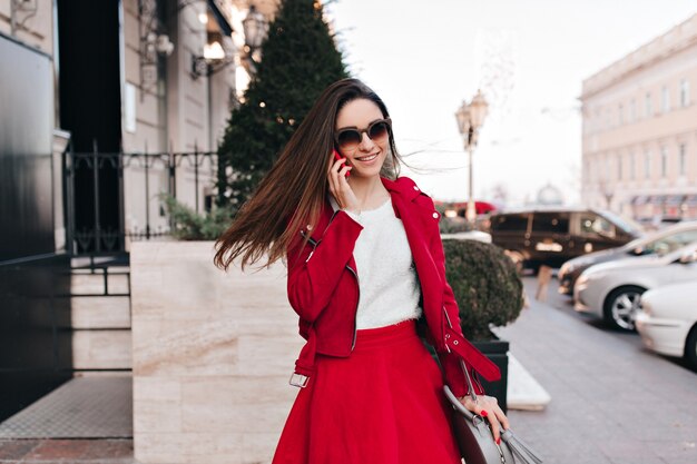 Pleased young white female model in red attire talking on phone
