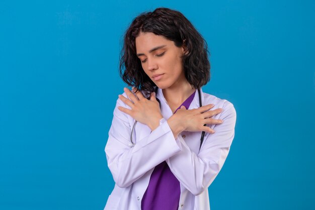 Pleased young woman doctor wearing white coat and with stethoscope standing with hands crossed on chest gesture feeling positive emotions on isolated blue